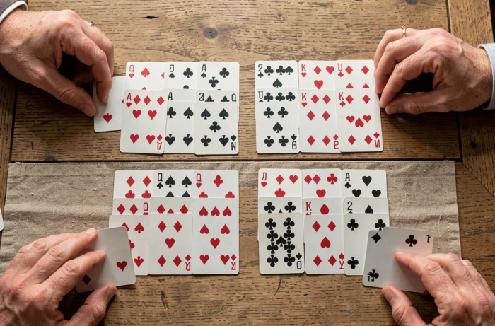 Vue du dessus d'une partie de Kems en cours avec des cartes réparties sur une table en chêne et les mains des joueurs visibles